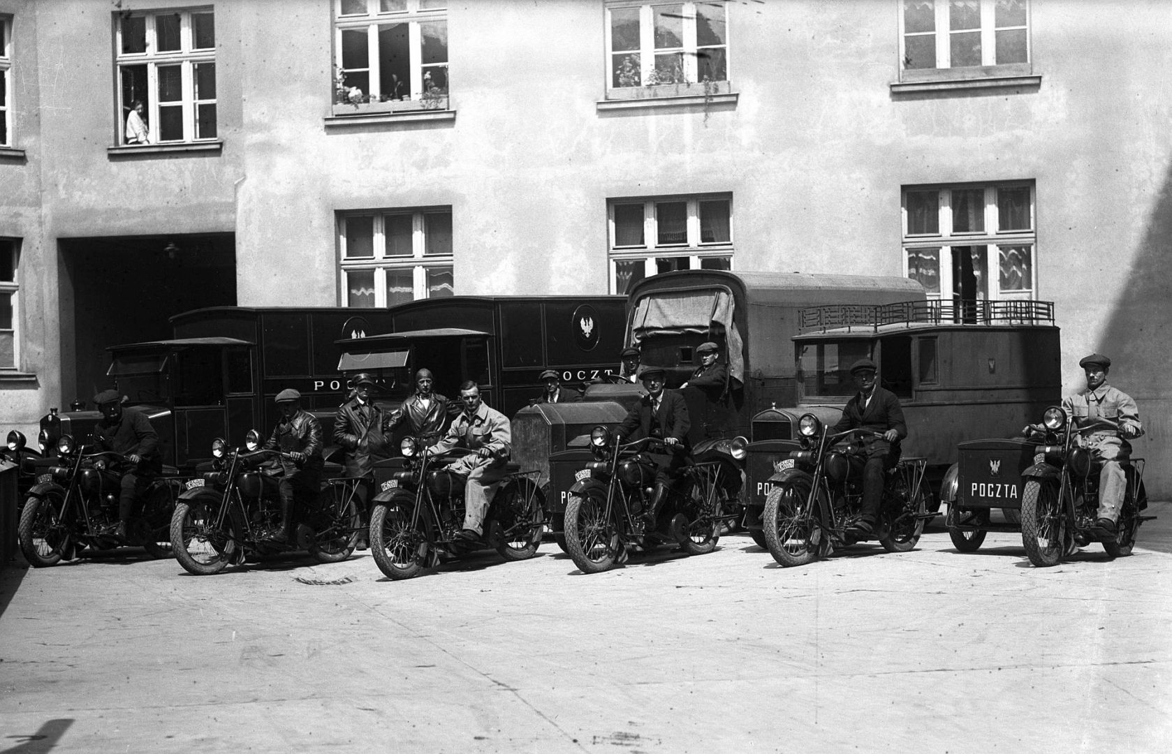 Mężczyźni na motocyklach, którzy zajmują się wybieraniem korespondencji ze skrzynek pocztowych. Z tyłu widoczne samochody pocztowe.